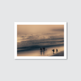 Photograph of surfers on Muriwai Beach, New Zealand, leaving the water and walking on the beach at either sunrise or sunset, with a moody and eery atmosphere.